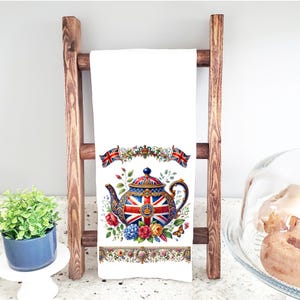 May include: A white tea towel with a colorful teapot design. The teapot is decorated with the Union Jack flag and a crown. The teapot is surrounded by flowers and butterflies.