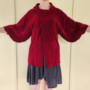 May include: A red velvet jacket with a wide collar and long sleeves. The jacket has a wrap-around style and is worn over a dark gray skirt.