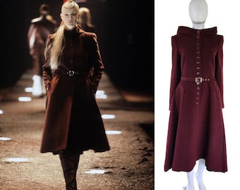 May include: A long, burgundy wool coat with a high collar, a button closure, and a belt. The coat has a flared skirt and is shown on a mannequin.
