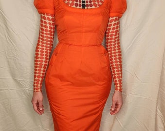 May include: A bright orange dress with a fitted bodice and a long skirt. The dress has a square neckline and puffed sleeves. The model is wearing a sheer top with a red and black polka dot pattern underneath the dress.