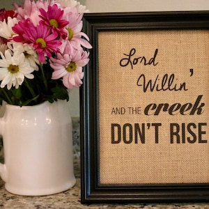 May include: A framed burlap print with the text "Lord Willin' and the creek don't rise" in black lettering. The print is in a black frame and is sitting on a table with a white pitcher of pink and white flowers.