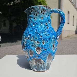 May include: A ceramic pitcher with a textured, turquoise blue glaze. The pitcher has a handle and a spout. The surface is decorated with dark brown and white spots. The pitcher is sitting on a white surface.