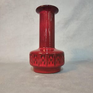Könnte beinhalten: Eine hohe, rote Keramikvase mit glänzendem Finish. Die Vase hat einen zylindrischen Hals und einen breiteren Sockel mit einem dekorativen Muster. Die Vase ist etwa 25 cm hoch und eignet sich perfekt zum Präsentieren von Blumen oder als dekoratives Element.
