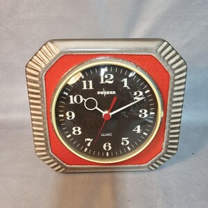 Könnte beinhalten: Eine quadratische Uhr in Rot und Silber mit schwarzen Zeigern und einem schwarzen Zifferblatt. Das Zifferblatt der Uhr hat weiße Zahlen und die Wörter "DEWETA" und "QUARTZ".