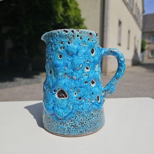 May include: A turquoise ceramic pitcher with a textured, bubbly surface and a handle. The pitcher has dark brown spots and a speckled base. The design is reminiscent of lava rock. The pitcher is likely used for serving beverages.