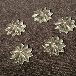 Op de afbeelding: Vijf heldere plastic bloemvormige versieringen met een stervormig ontwerp. De versieringen zijn ongeveer 2,5 cm in diameter.