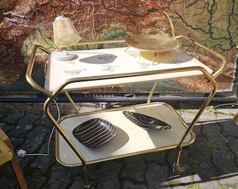 May include: A gold metal serving cart with two tiers. The top tier has a white surface with a floral pattern and a brass serving dish. The bottom tier has a white surface with two black and gold bowls. The cart has wheels and is on a brick surface.