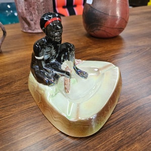 May include: A ceramic ashtray featuring a figurine of a person sitting on a leaf-shaped base. The person is depicted with dark skin, black hair, and a red headband. The ashtray is decorated with a green and brown glaze.