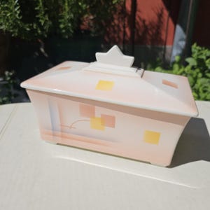 Könnte beinhalten: Rechteckige Keramikbox mit Deckel in Rosa-, Pfirsich- und Gelbtönen. Die Box hat einen sternförmigen Griff und geometrische Muster. Wahrscheinlich für die Aufbewahrung oder Präsentation gedacht.