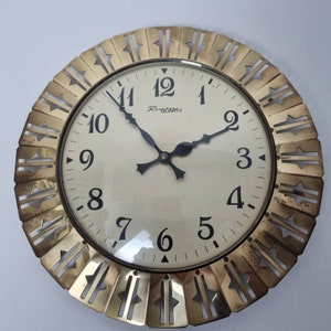 Puede incluir: Un reloj de pared de color dorado con una esfera blanca y marcadores de hora en números romanos negros. El reloj tiene un marco decorativo y ornamentado con un patrón repetitivo de estrellas y formas geométricas. La esfera del reloj dice "Richtartz".