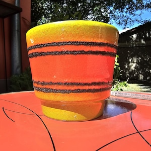 Può includere: Un vaso di ceramica dal design vivace. Le sezioni superiore e inferiore sono gialle, mentre la sezione centrale è arancione. Tre bande nere circondano la sezione arancione. Il vaso poggia su una superficie rossa.