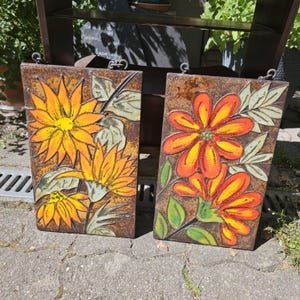 Puede incluir: Dos baldosas de cerámica rectangulares con diseños florales. Una baldosa presenta girasoles amarillos, la otra flores naranjas y amarillas. Ambas baldosas tienen un fondo marrón y hojas verdes. Cada baldosa tiene un lazo metálico para colgar.