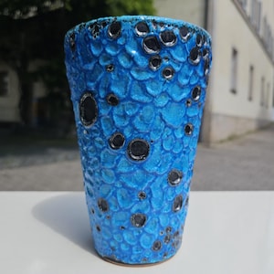 May include: A tall, cylindrical ceramic vase with a textured, scale-like pattern in shades of bright blue. The vase features irregular, dark gray circular accents. The vase is approximately 8 inches tall.