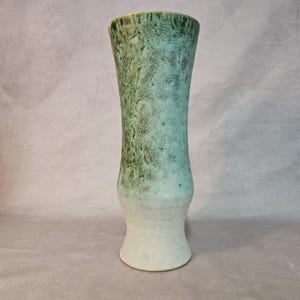 Könnte beinhalten: Eine hohe Keramikvase mit strukturierter Oberfläche. Die Vase hat einen Farbverlauf, der von einem weißen Sockel über eine hellblaue Mitte zu einer dunkelgrünen Oberseite verläuft. Die Vase hat einen ausgestellten oberen Rand und einen abgerundeten Sockel.