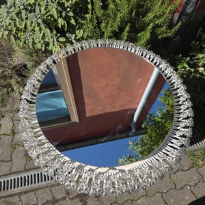 Op de afbeelding: Een ronde spiegel met een decoratieve, stervormige rand. De spiegel reflecteert een blauwe lucht en delen van een gebouw. De rand is gemaakt van een zilverkleurig materiaal, wat een vleugje elegantie aan het ontwerp toevoegt.