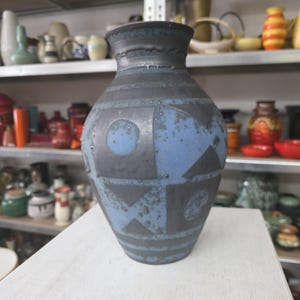 Könnte beinhalten: Eine Keramikvase in Dunkelblau und Grau. Die Vase hat geometrische Formen und horizontale Streifen. Die Vase steht auf einer weißen Oberfläche, mit Regalen mit anderer Keramik im Hintergrund.