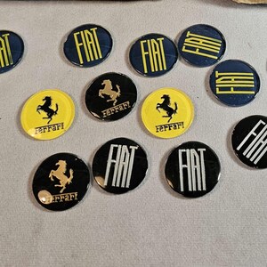 Può includere: Assortimento di adesivi rotondi con logo di auto di vari marchi, tra cui Fiat, Ferrari e BMW. Gli adesivi sono neri, bianchi, gialli e blu con lettere bianche.