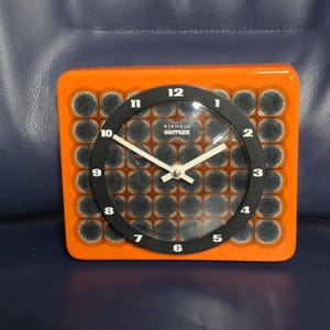 Könnte beinhalten: Eine orangefarbene rechteckige Kienzle Boutique Uhr mit schwarzem Zifferblatt und weißen Zeigern. Das Zifferblatt hat schwarze Zahlen und den Text "Kienzle Boutique". Der Hintergrund hat ein Muster aus dunkelgrauen Kreisen.