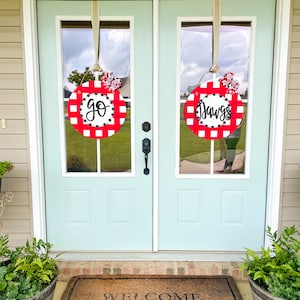 May include: Two round, red and white checkered door hangers with the words "go" and "Dawgs" in black script. Each hanger has a decorative bow and is suspended from a light blue door.