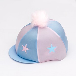 K&ouml;nnte beinhalten: Ein Reithelm in Pastellfarben mit hellblauem Schirm und einem flauschigen rosa Bommel. Der Helm ist in hellblaue und lavendelfarbene Abschnitte unterteilt, die jeweils mit sternf&ouml;rmigen Akzenten in Rosa und Hellblau verziert sind.