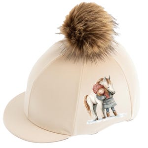 May include: A beige equestrian helmet with a brown faux fur pom-pom on top. The helmet features a graphic of a girl hugging a white horse. This riding helmet is designed for equestrian sports.