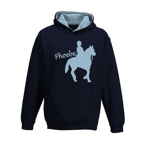 Può includere: Felpa con cappuccio blu navy con interno cappuccio azzurro e tasca a marsupio. La parte anteriore presenta una silhouette azzurra di una persona a cavallo, con il nome "Phoebe" in corsivo.