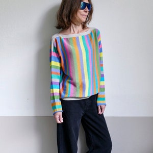 Puede incluir: Un suéter a rayas multicolores con cuello barco. El suéter presenta rayas verticales en varios colores, incluyendo rosa, amarillo, verde y azul. La persona lleva pantalones de pana negros y zapatillas deportivas azules.