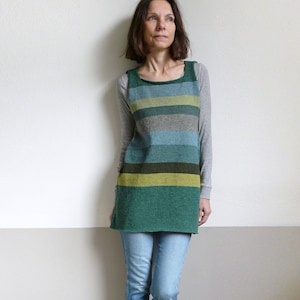 May include: A woman wearing a green, blue, and gray striped tunic over a gray long-sleeved shirt, paired with blue jeans and gray sneakers. The tunic has a square neckline and a straight silhouette, with horizontal stripes in varying shades of green, blue, and gray.