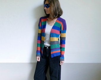 Op de afbeelding: Een vrouw draagt een gestreepte cardigan met een wit tanktop eronder en een donkerblauwe jeans.