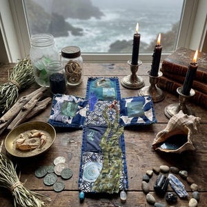 May include: A cross-shaped fabric textile featuring blue and white patterned patches, green fabric strips, and small images. It is surrounded by glass jars, black candles in silver holders, dried grass, driftwood, coins, a crab, and stones.