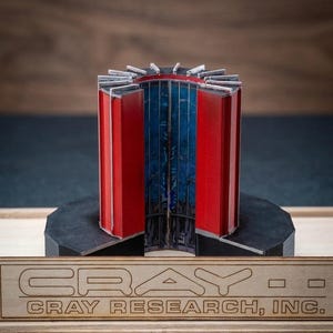 May include: A model of a Cray Research supercomputer component. The model features a cylindrical design with red and blue panels, mounted on a black base. The base rests on a wooden platform with the "CRAY RESEARCH, INC." logo.