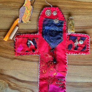 May include: A handmade red felt cross-shaped item with white stitching, pockets, and decorative charms. A blue rose print is visible. Additional items include a paper bird, candles, and a small glass bottle.