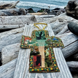 May include: A cross-shaped fabric wall hanging featuring a collage of printed images including a tree, a fairy, and a red door. The border is decorated with a yellow floral pattern on a dark brown background, and a yellow ribbon loop is attached to the top.