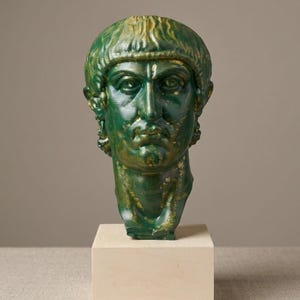 May include: A green and gold painted bust of a person with short, straight hair and a serious expression. The sculpture features detailed facial features, including defined eyes, a straight nose, and a firm mouth, mounted on a square wooden base.