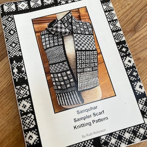 Puede incluir: Un patrón de tejido titulado "Sanquhar Sampler Scarf Knitting Pattern" de Ruth Robinson. La portada presenta una bufanda con estampado en blanco y negro sobre un maniquí. El libro tiene un borde blanco con un diseño geométrico en blanco y negro.