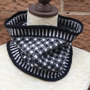 May include: A black and white knitted cowl scarf. The scarf features a checkered pattern in the center and striped borders. The cowl is displayed on a mannequin, showcasing its design and texture. The color scheme is classic and versatile.