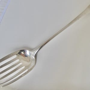 May include: A large, silver serving fork with a long, slender handle and a wide head featuring six tines. The fork is highly polished, reflecting the surrounding environment. The handle has a rectangular end. The utensil is designed for serving food.