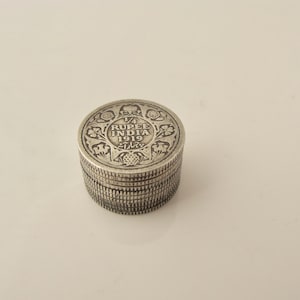 May include: A small, round, silver-colored box with intricate floral and geometric designs. The top of the box features the text "1/4 RUPEE INDIA 1919" in a circular arrangement. The box has a ribbed side and a slightly domed lid.