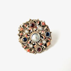 Op de afbeelding: Een vintage, ronde broche met een goudkleurig metalen frame. De broche heeft een centrale iriserende parel, kleinere parels, rode en blauwe edelstenen en koraalkleurige emaille accenten. Het ontwerp is sierlijk en gedetailleerd.