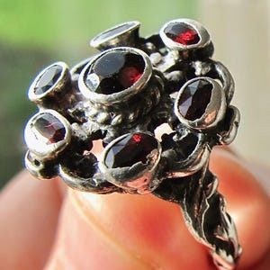 Op de afbeelding: Een zilveren ring met meerdere ovaal geslepen, dieprode edelstenen. De ring heeft een textuur, organisch ontwerp met de edelstenen in individuele bezels. De ring wordt vastgehouden door de vingers van een persoon.