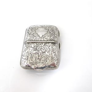 May include: A silver rectangular case with intricate floral engravings. The case has a hinged lid and a clasp closure. The surface is highly detailed with swirling patterns and a small, blank cartouche on the top. The case is likely antique.