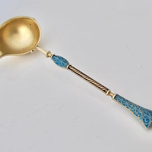 Pode incluir: Uma concha dourada com uma tigela arredondada e um cabo longo. O cabo apresenta detalhes em esmalte azul com um padrão decorativo e uma seção central dourada torcida. A concha é provavelmente para servir molhos ou sopas.