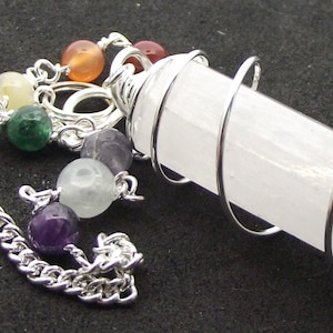 Op de afbeelding: Een witte kristallen slinger met een zilveren draad eromheen gewikkeld. De slinger is bevestigd aan een zilveren ketting met zeven kralen in verschillende kleuren, waaronder helder, geel, groen, oranje, paars en lichtgroen.