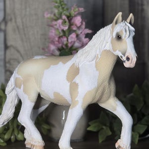 May include: A plastic horse figurine featuring a cream and white pinto coat pattern. The horse has a textured white mane and tail, with detailed hooves and a realistic facial expression, standing in a poised position with one front leg lifted.