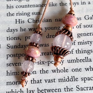 May include: Two handmade beaded bookmarks with pink and clear gemstone beads and copper-coloured metal accents. The bookmarks are hanging over a book page with printed text.