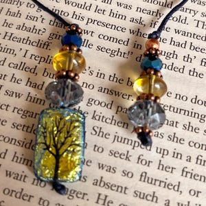 May include: Two handmade tree pendant necklaces. Each necklace features a rectangular pendant with a tree design in black, yellow, and blue. The pendants are strung with beads in blue, yellow, grey, and copper tones.