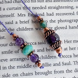 May include: Two handmade beaded bookmarks. Each bookmark features a lilac cord, amethyst, turquoise, and copper-coloured beads. The beads are arranged in a decorative pattern, creating a unique and artistic design. Ideal for book lovers.