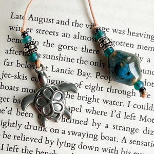 May include: A silver turtle charm on a beaded necklace with turquoise and silver beads. The necklace is on a book with the text "Late August and the village was heaving" visible.