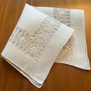 May include: A white square handkerchief featuring a wide border of intricate needle lace with floral and geometric patterns. The fabric has a visible woven texture, and the lace insert creates a decorative frame around the central linen panel.