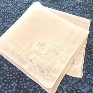 May include: Off-white handkerchief with a geometric embroidered design. The handkerchief is folded, revealing the detailed stitching along the edges and the decorative pattern. The background is a dark blue fabric with a subtle pattern.